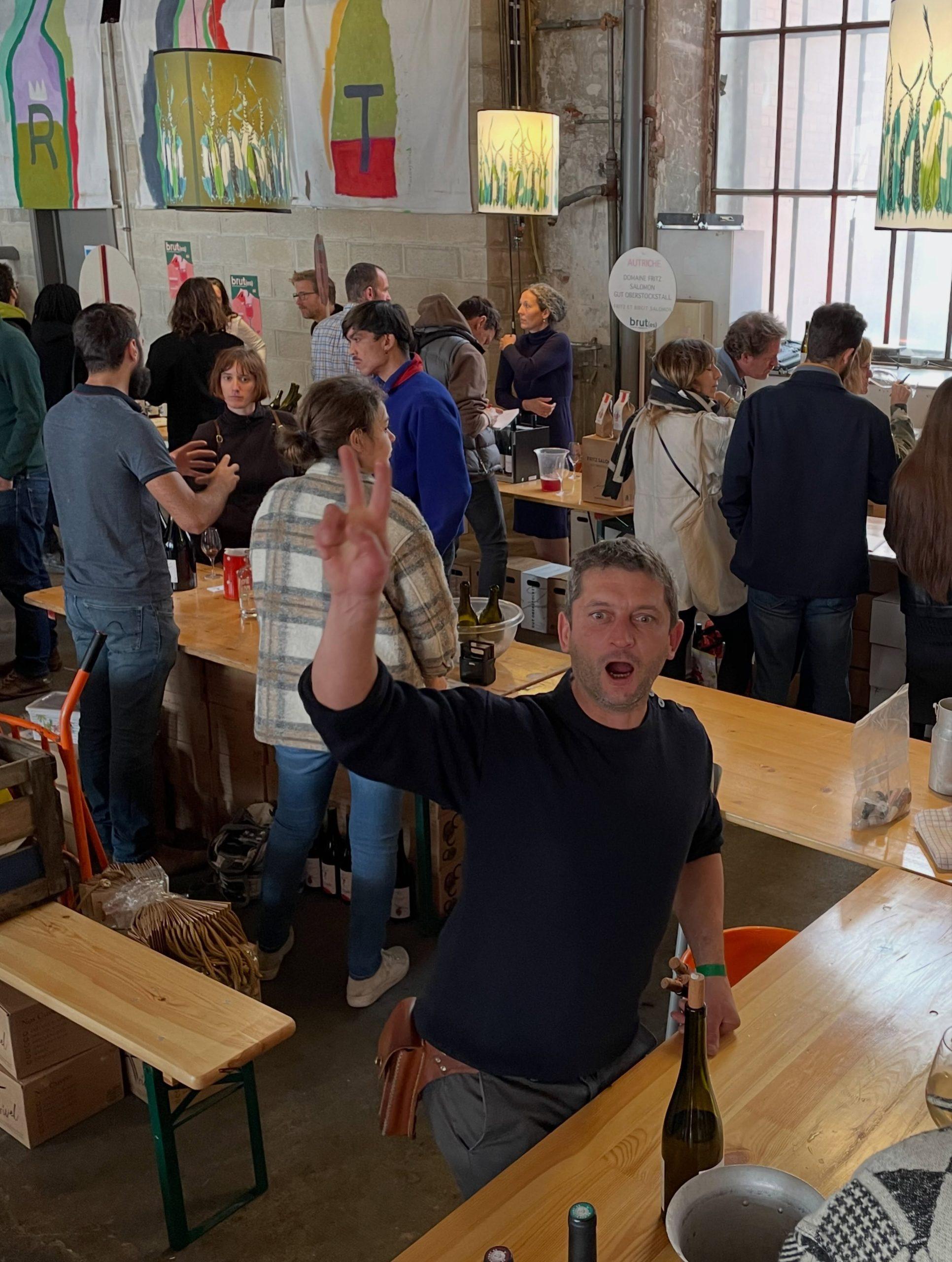 Un exposant fait le signe de la victoire derrière les tables de dégustation du salon Brut(es), au milieu des visiteurs